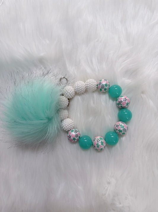 A beaded wristlet with a fluffy mint green pom-pom charm, The bracelet features white textured beads, solid mint green beads, and floral-patterned beads with pink, green, and white colors.