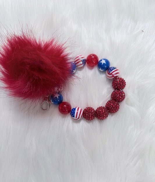 A patriotic-themed beaded bracelet with a large red faux fur pom-pom charm. The bracelet features red glitter beads, red and white striped beads, and blue beads with white stars